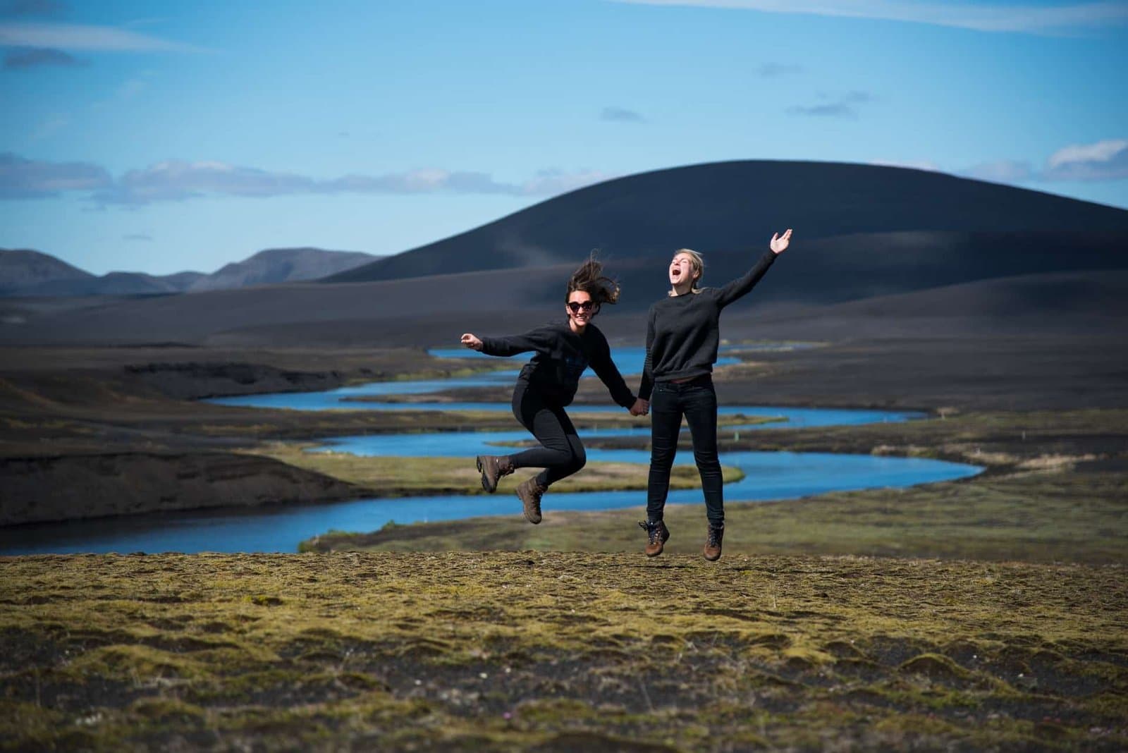 Two people jumping joyfully on a hillside above a winding Icelandic river